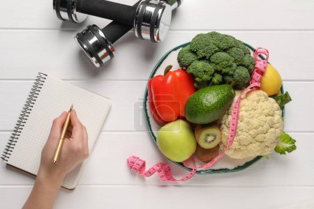 Woman developing diet plan at white wooden table with products and dumbbells, top view