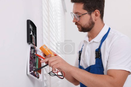 Technician using digital multimeter while installing home security alarm system on white wall indoors