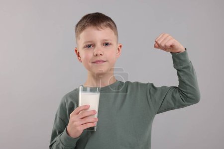 Téléchargez les photos : Mignon garçon avec verre de lait frais montrant sa force sur fond gris clair - en image libre de droit