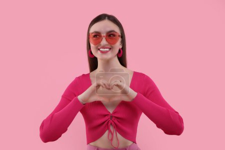 Pink look. Beautiful woman in sunglasses making heart with hands on color background