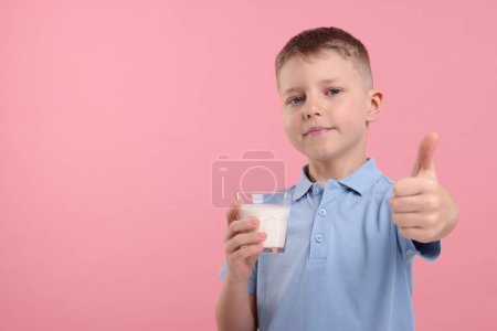 Téléchargez les photos : Garçon mignon avec verre de lait frais montrant pouce vers le haut sur fond rose, espace pour le texte - en image libre de droit