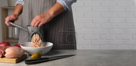 Woman making chicken mince with manual meat grinder at grey table, closeup. Banner design with space for text
