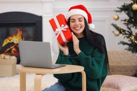 Celebrando la Navidad en línea con regalos intercambiados por correo. Mujer sonriente en sombrero de Santa con caja de regalo durante la videollamada en el ordenador portátil en casa