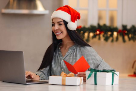 Foto de Celebrando la Navidad en línea con regalos intercambiados por correo. Mujer sonriente en el sombrero de Santa con tarjeta de felicitación y regalos durante la videollamada en casa - Imagen libre de derechos