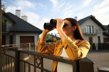 Téléchargez les photos : Concept de vie privée. Curieux jeune femme avec des jumelles espionnant les voisins sur la clôture à l'extérieur - en image libre de droit