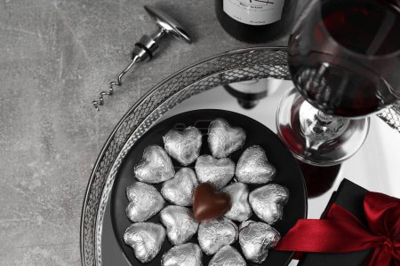 Bottle, glass of red wine, heart shaped chocolate candies, corkscrew and gift box on light grey textured table, above view. Space for text