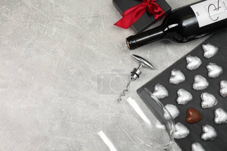 Bottle of red wine, glasses, heart shaped chocolate candies, corkscrew and gift box on light grey textured table, flat lay. Space for text