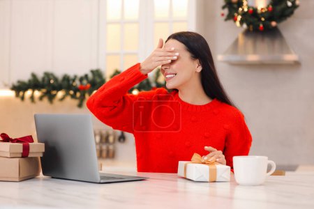 Weihnachten online feiern und Geschenke per Post austauschen. Lächelnde Frau verdeckt Augen, bevor sie während eines Videotelefonats zu Hause die Geschenkbox öffnet