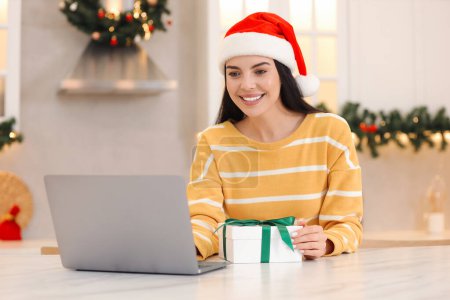 Celebrating Christmas online with exchanged by mail presents. Smiling woman in Santa hat with gift box during video call on laptop at home