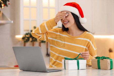 Celebrating Christmas online with exchanged by mail presents. Smiling woman covering eyes before opening gift box during video call at home