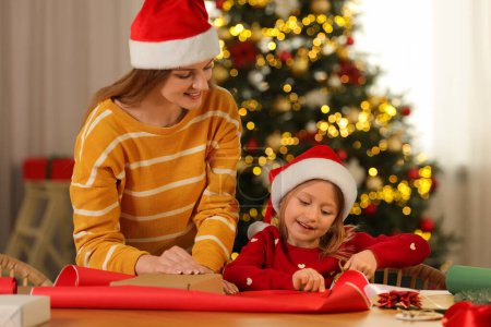 Mother and her little daughter in Santa hats wrapping Christmas gift at home