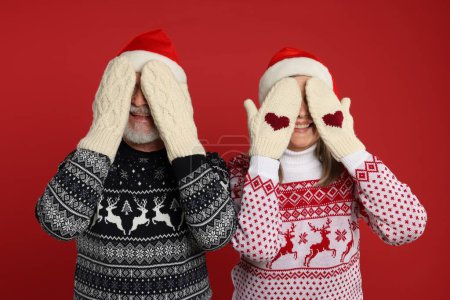 Senior couple in Christmas sweaters and Santa hats covering faces with hands in knitted mittens on red background