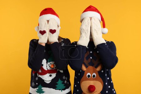Couple in Christmas sweaters and Santa hats covering faces with hands in knitted mittens on orange background