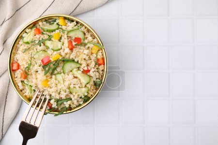 Cooked bulgur with vegetables in bowl on white tiled table, top view. Space for text