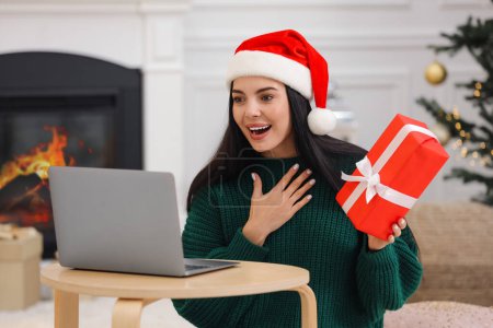 Celebrando la Navidad en línea con regalos intercambiados por correo. Mujer sorprendida en Santa sombrero agradeciendo el regalo durante la videollamada en casa