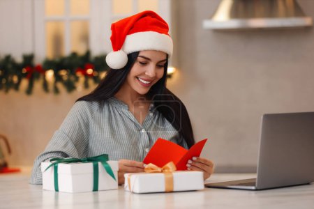 Celebrando la Navidad en línea con regalos intercambiados por correo. Mujer sonriente en el sombrero de Santa con tarjeta de felicitación y regalos durante la videollamada en casa