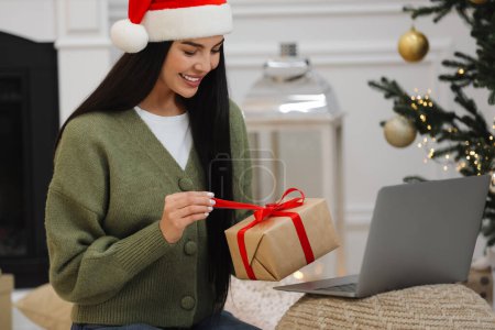 Celebrando la Navidad en línea con regalos intercambiados por correo. Mujer sonriente abriendo caja de regalo durante la videollamada en el ordenador portátil en casa