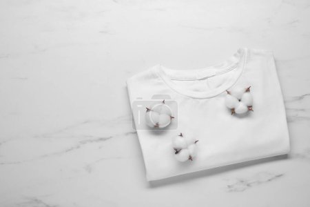 Camiseta y flores de algodón esponjoso sobre fondo de mármol blanco, vista superior. Espacio para texto