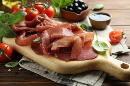 Photo for Board with delicious bresaola served with tomato and basil leaves on wooden table, closeup - Royalty Free Image