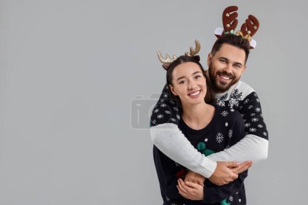Feliz pareja joven en suéteres de Navidad y diademas de renos sobre fondo gris. Espacio para texto
