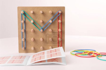 Wooden geoboard with letter M made of rubber bands and instruction on white table. Educational toy for motor skills development