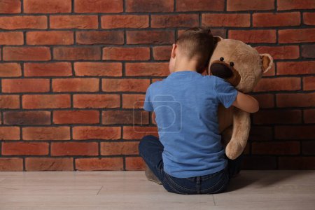 Child abuse. Upset boy with teddy bear sitting on floor near brick wall indoors, back view. Space for text