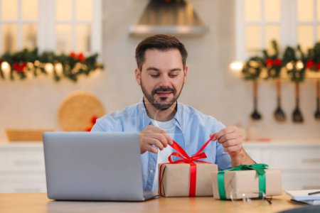 Celebrating Christmas online with exchanged by mail presents. Man opening gift box during video call on laptop at home
