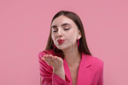 Beautiful woman in pink clothes blowing kiss on color background