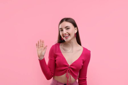 Beautiful woman in pink clothes waving hello on color background