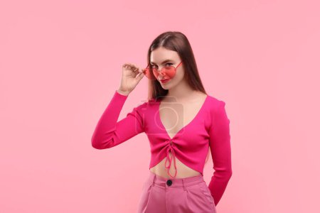 Beautiful woman in pink clothes and heart shaped sunglasses on color background