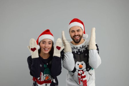 Jeune couple en pulls de Noël, chapeaux de Père Noël et mitaines tricotées sur fond gris