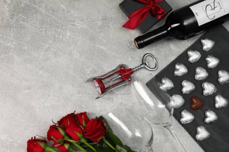 Bottle of red wine, glasses, heart shaped chocolate candies, corkscrew, roses and gift box on light grey textured table, flat lay. Space for text