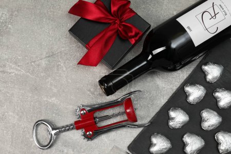 Bottle of red wine, heart shaped chocolate candies, corkscrew and gift box on light grey textured table, flat lay