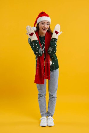 Happy young woman in Christmas sweater, Santa hat and knitted mittens on orange background
