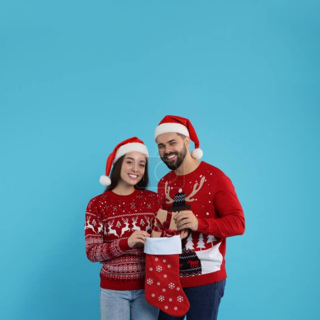 Glückliches junges Paar in Weihnachtspullis und Nikolausmützen, die Geschenke aus dem Strumpf auf hellblauem Hintergrund entgegennehmen
