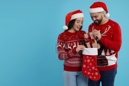 Glückliches junges Paar in Weihnachtspullis und Nikolausmützen, die Geschenke aus dem Strumpf auf hellblauem Hintergrund entgegennehmen. Raum für Text