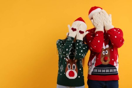 Pareja en suéteres de Navidad y sombreros de Santa cubriendo caras con las manos en mitones de punto sobre fondo naranja. Espacio para texto