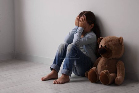 Child abuse. Upset little girl with teddy bear sitting on floor near light wall indoors, space for text