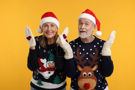 Feliz pareja de ancianos en suéteres de Navidad, sombreros de Santa Claus y mitones de punto sobre fondo naranja