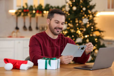 Foto de Celebrando la Navidad en línea con regalos intercambiados por correo. Feliz hombre leyendo la tarjeta de felicitación durante la videollamada en el ordenador portátil en casa - Imagen libre de derechos