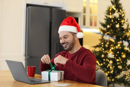 Weihnachten online feiern und Geschenke per Post austauschen. Glücklicher Mann mit Weihnachtsmütze öffnet Geschenkbox während Videoanruf auf Laptop zu Hause