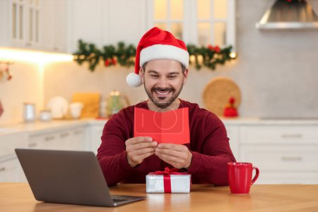 Celebrating Christmas online with exchanged by mail presents. Happy man reading greeting card during video call on laptop at home