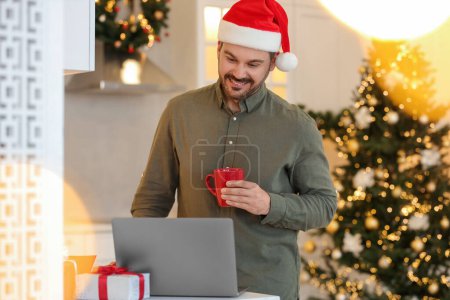 Celebrando la Navidad en línea con regalos intercambiados por correo. Hombre feliz en el sombrero de Santa con taza de bebida durante la videollamada en el ordenador portátil en casa