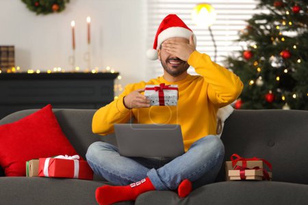 Celebrando la Navidad en línea con regalos intercambiados por correo. Hombre en Santa sombrero cubriendo los ojos antes de abrir la caja de regalo durante la videollamada en el ordenador portátil en casa
