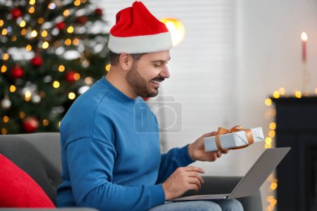 Célébrons Noël en ligne avec des cadeaux échangés par la poste. Homme heureux dans le chapeau de Père Noël avec boîte cadeau pendant l'appel vidéo sur ordinateur portable à la maison