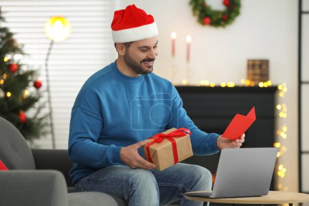 Celebrating Christmas online with exchanged by mail presents. Smiling man in Santa hat with greeting card and gift box during video call on laptop at home