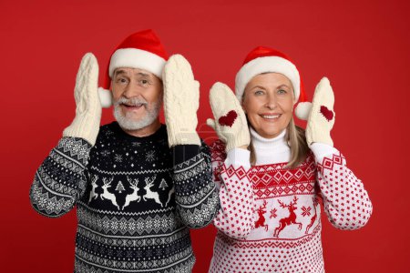Couple aîné en pulls de Noël, chapeaux de Père Noël et mitaines tricotées sur fond rouge
