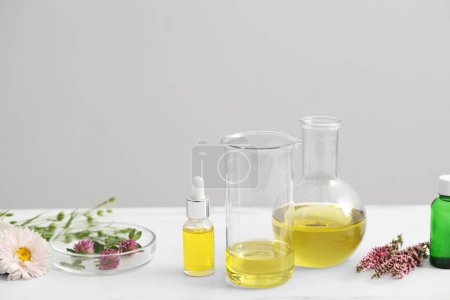 Cosmetic oil, laboratory dishware and flowers on white table