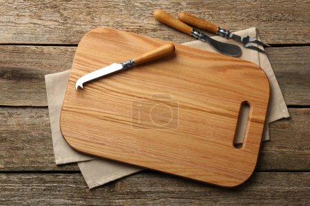 Cutting board, cheese knives and fork on old wooden table, flat lay