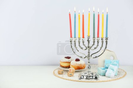 Chanukka-Feier. Menora mit brennenden Kerzen, Dreidels, Donuts und Geschenkschachteln auf weißem Holztisch, Platz für Text